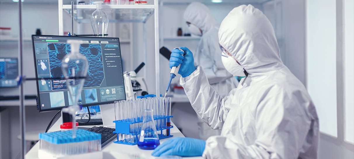 Wissenschaftler im Schutzanzug, der in einem Reinraum eine Mikropipette an einem Reagenzglas verwendet. Im Hintergrund ist ein weiterer Mitarbeiter zu sehen.