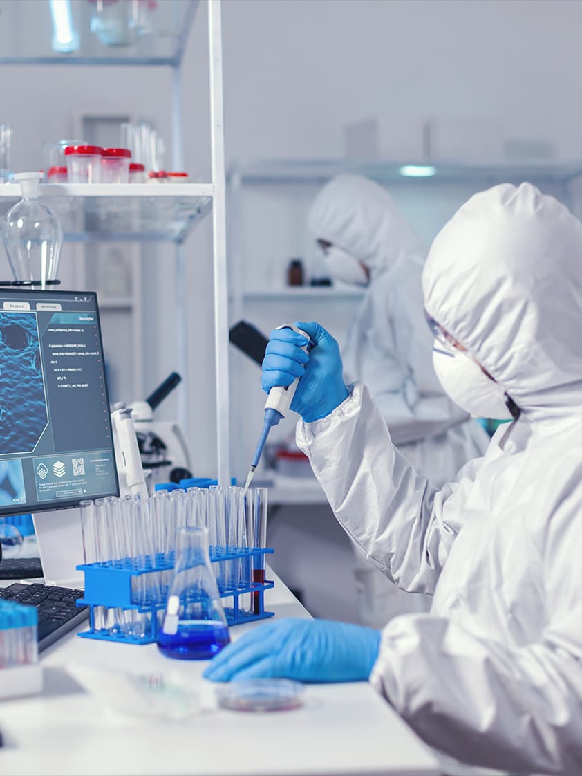 Wissenschaftler im Schutzanzug, der in einem Reinraum eine Mikropipette an einem Reagenzglas verwendet. Im Hintergrund ist ein weiterer Mitarbeiter zu sehen.